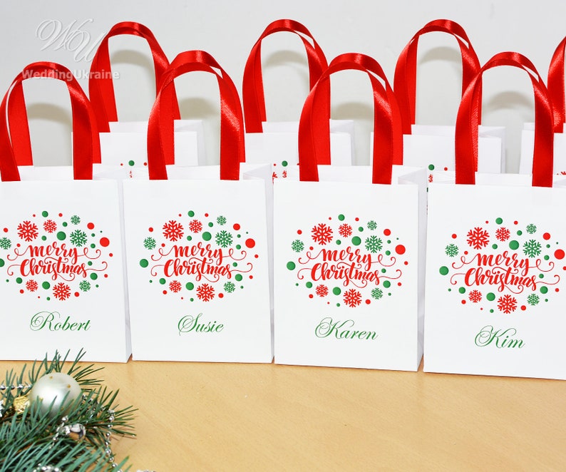 May include: Four white gift bags with red handles and a green and red snowflake design. The bags have the words 'merry christmas' printed on them and each bag has a name printed on the bottom: Robert, Susie, Karen, and Kim.
