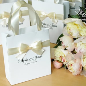 Puede incluir: Bolsas de regalo blancas con lazos de cinta de sat&eacute;n marfil y el texto "Nycha & Junior August 18, 2016". Las bolsas est&aacute;n dispuestas sobre una mesa de madera con un ramo de peon&iacute;as rosa p&aacute;lido.