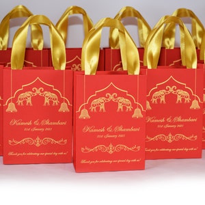 Op de afbeelding: Rode en gouden cadeauzakjes met gouden lintjes. De zakjes hebben een ontwerp met twee olifanten met bellen en de tekst "Kamesh & Shambavi 31st January 2021" en "Bedankt dat je onze speciale dag met ons hebt gevierd!"