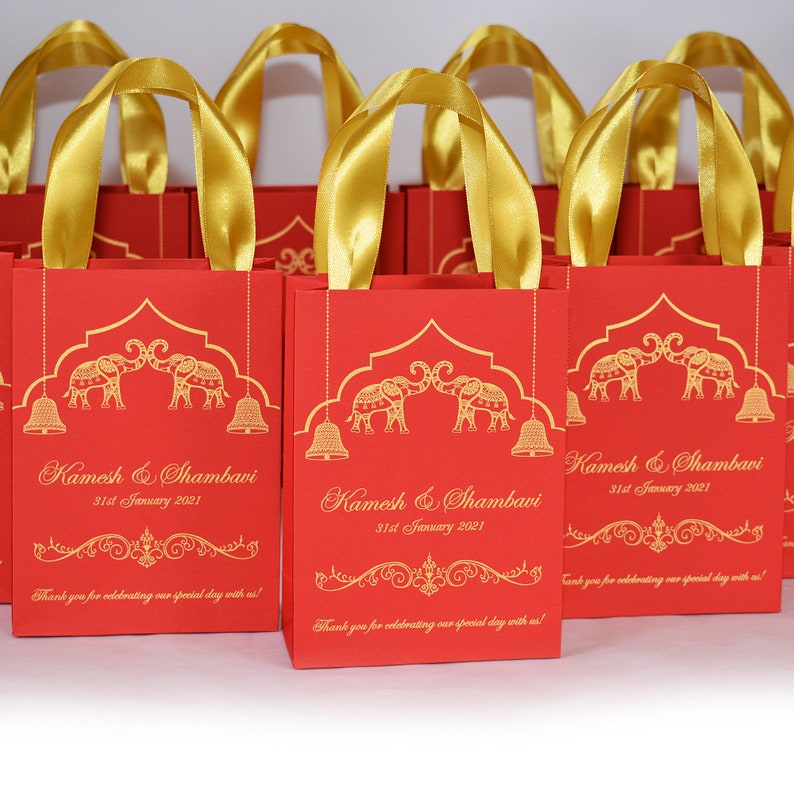 Op de afbeelding: Rode en gouden cadeauzakjes met gouden lintjes. De zakjes hebben een ontwerp met twee olifanten met bellen en de tekst "Kamesh & Shambavi 31st January 2021" en "Bedankt dat je onze speciale dag met ons hebt gevierd!"