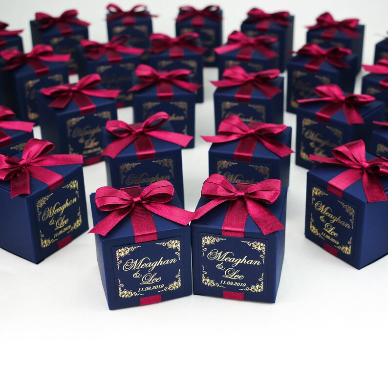 May include: A collection of navy blue gift boxes, each adorned with a burgundy ribbon bow. The boxes feature gold labels with text. They are presented on a white surface, likely for a special occasion.