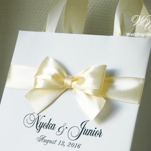 Puede incluir: Una bolsa de regalo blanca con un lazo de cinta de sat&eacute;n amarillo y el texto "Nyoka & Junior August 13, 2016".