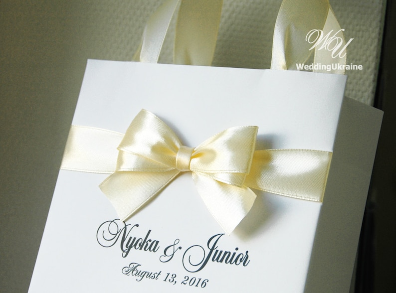 Puede incluir: Una bolsa de regalo blanca con un lazo de cinta de sat&eacute;n amarillo y el texto "Nyoka & Junior August 13, 2016".