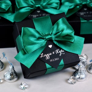 May include: A black favor box with a green satin ribbon tied in a bow. The box has a black tag with white text that reads "Lizzie & Kyle 10.2.2021".
