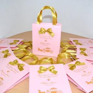 May include: Pink and gold favor bags with gold ribbon handles and bows. The bags have a gold crown and carriage design with the text "Thank you for welcoming our sweet little princess, Skylar Threne, October 2019".