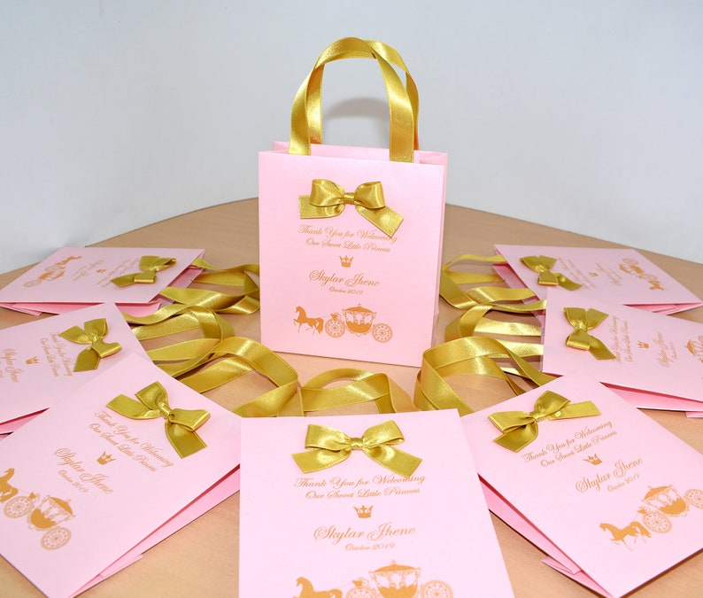 May include: Pink and gold favor bags with gold ribbon handles and bows. The bags have a gold crown and carriage design with the text "Thank you for welcoming our sweet little princess, Skylar Threne, October 2019".