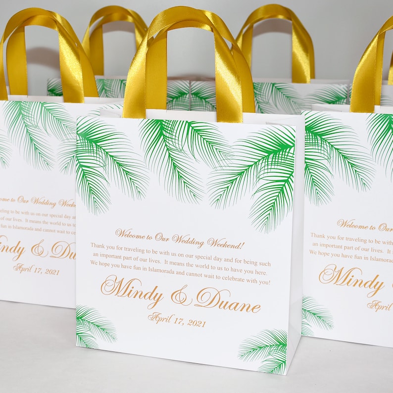 May include: White favor bags with green palm leaf designs and gold ribbon handles. The bags have a welcome message for a wedding weekend with the names Mindy & Duane and the date April 17, 2021.