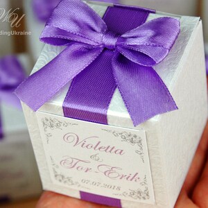 May include: A white favor box with a purple satin ribbon and bow. The box has a label with the text "Violetta & Tor-Erik 07.07.2018".