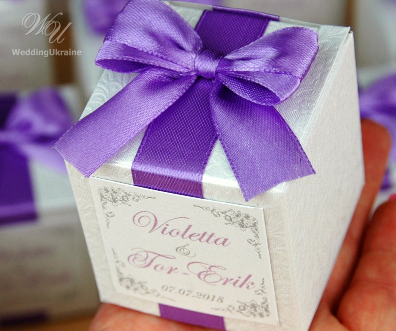 May include: A white favor box with a purple satin ribbon and bow. The box has a label with the text "Violetta & Tor-Erik 07.07.2018".
