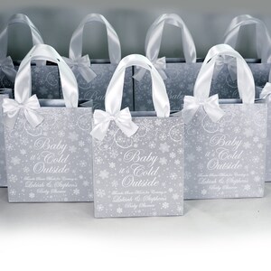 May include: A set of silver gift bags with white satin ribbon handles and bows. The bags are decorated with a snowflake pattern and the text "Baby it's Cold Outside" with the names Lakiah & Stephen's Baby Shower.