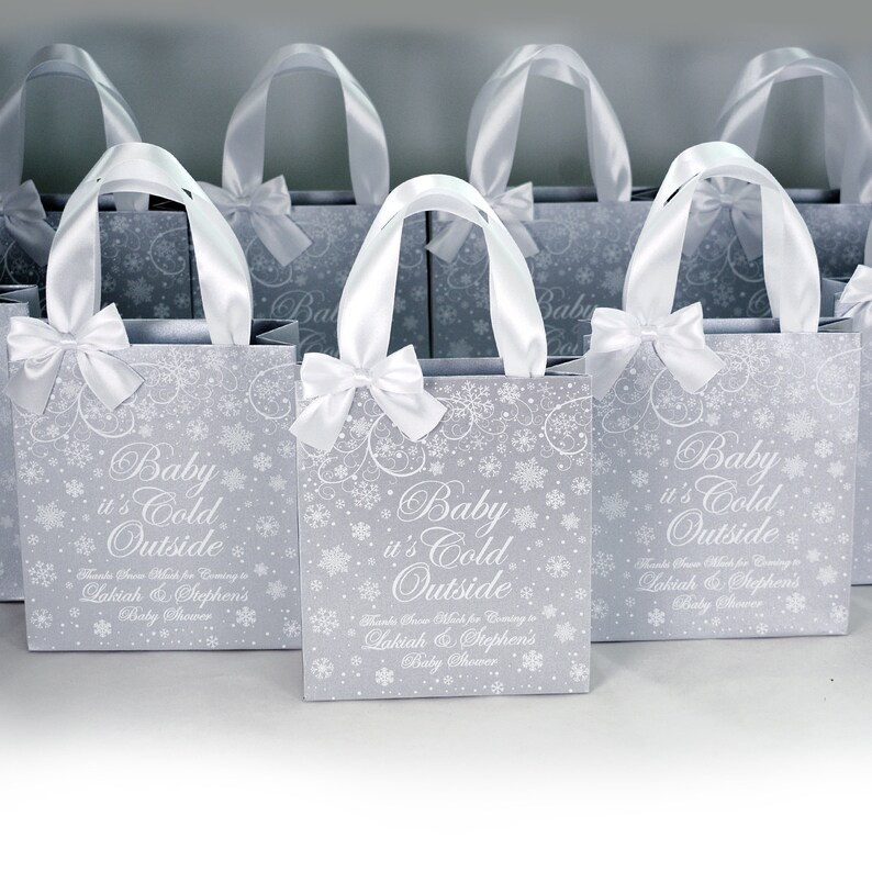 May include: A set of silver gift bags with white satin ribbon handles and bows. The bags are decorated with a snowflake pattern and the text "Baby it's Cold Outside" with the names Lakiah & Stephen's Baby Shower.