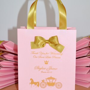 May include: Pink favor bag with gold handles and bow. The bag has a gold crown and carriage design with the text "Thank You for Welcoming Our Sweet Little Princess Skylar Jhene October 2019".