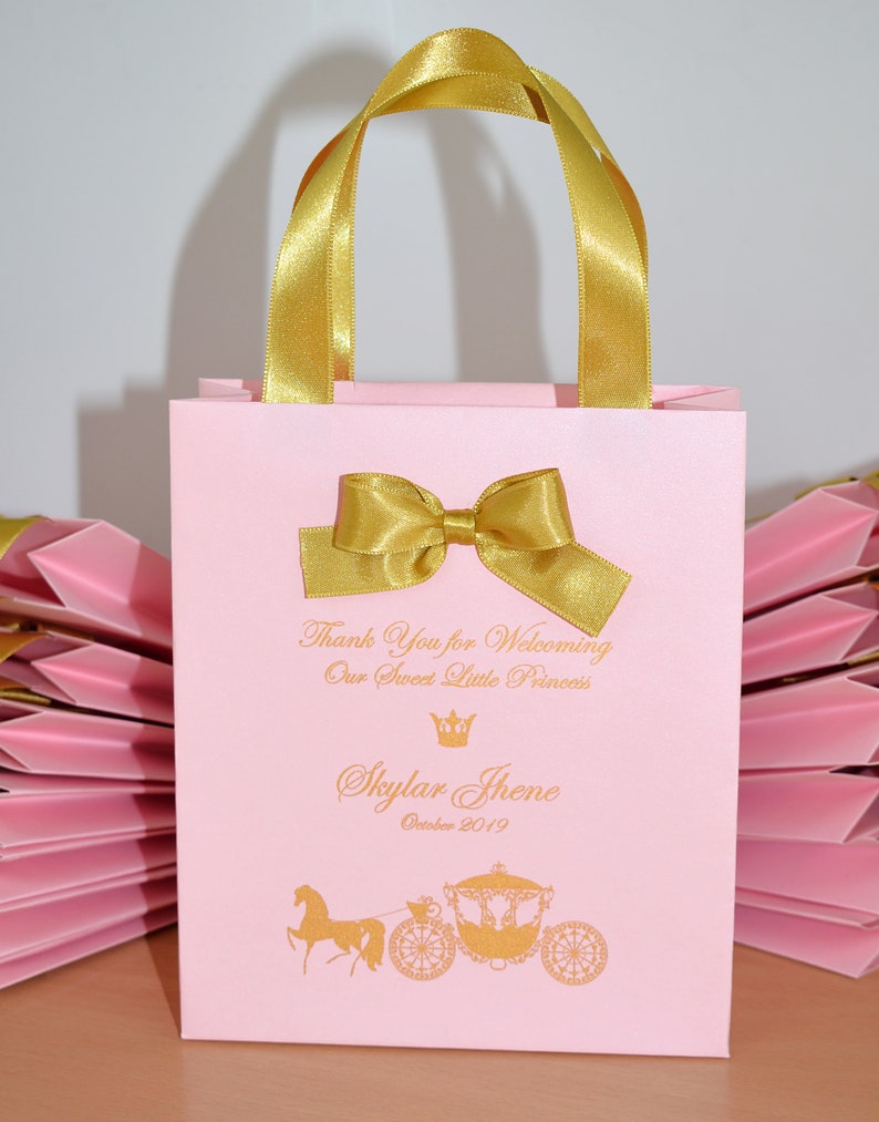 May include: Pink favor bag with gold handles and bow. The bag has a gold crown and carriage design with the text "Thank You for Welcoming Our Sweet Little Princess Skylar Jhene October 2019".