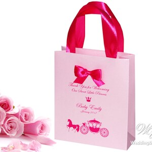 May include: Pink gift bag with a pink ribbon and bow. The bag has a printed design of a carriage and horse, a crown, and the text "Thank you for welcoming our sweet little princess. Baby Emily January 2017".
