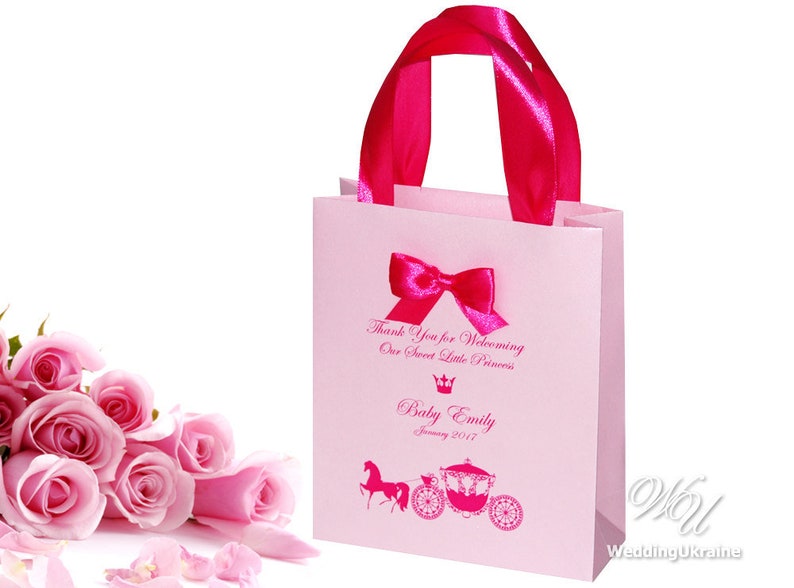 May include: Pink gift bag with a pink ribbon and bow. The bag has a printed design of a carriage and horse, a crown, and the text "Thank you for welcoming our sweet little princess. Baby Emily January 2017".