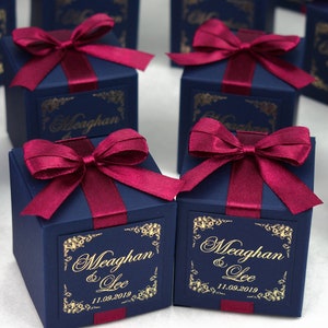 May include: Dark blue gift boxes tied with burgundy ribbon bows. Each box features a gold label with the names "Meaghan & Lee" and the date "11.09.2019". The boxes are displayed on a white surface, suitable for weddings or celebrations.
