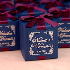May include: Navy blue gift boxes tied with burgundy ribbon and bows. Each box has a gold label with the names "Natasha & Dennis" and the date "4.28.2018". The boxes are on a light-coloured surface.