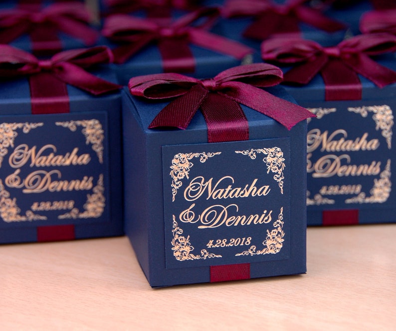 May include: Navy blue gift boxes tied with burgundy ribbon and bows. Each box has a gold label with the names "Natasha & Dennis" and the date "4.28.2018". The boxes are on a light-coloured surface.