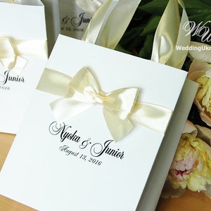 Puede incluir: Bolsas de regalo blancas con lazos de cinta de sat&eacute;n marfil y el texto "Nycka & Junior August 13, 2016" impreso en negro.