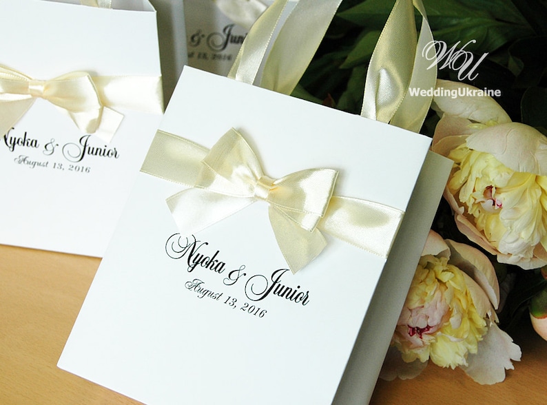Puede incluir: Bolsas de regalo blancas con lazos de cinta de sat&eacute;n marfil y el texto "Nycka & Junior August 13, 2016" impreso en negro.