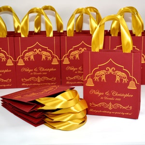 Op de afbeelding: Rode en gouden cadeauzakjes met gouden lintjes als handvatten. De zakjes hebben een ontwerp met twee olifanten, een decoratieve boog en hangende bellen. De zakjes zijn gepersonaliseerd met de namen "Nithya & Christopher" en de datum "30 december 2022".
