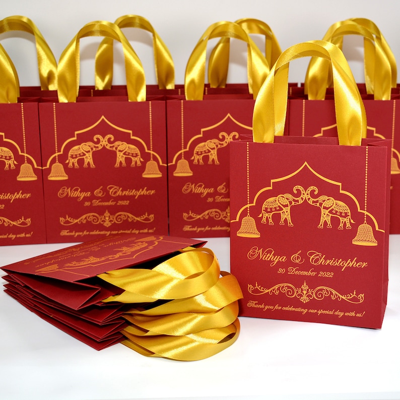 Op de afbeelding: Rode en gouden cadeauzakjes met gouden lintjes als handvatten. De zakjes hebben een ontwerp met twee olifanten, een decoratieve boog en hangende bellen. De zakjes zijn gepersonaliseerd met de namen "Nithya & Christopher" en de datum "30 december 2022".