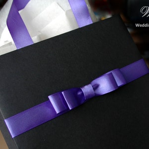 May include: A black gift bag with a purple satin ribbon tied in a bow. The bag is filled with white tissue paper.