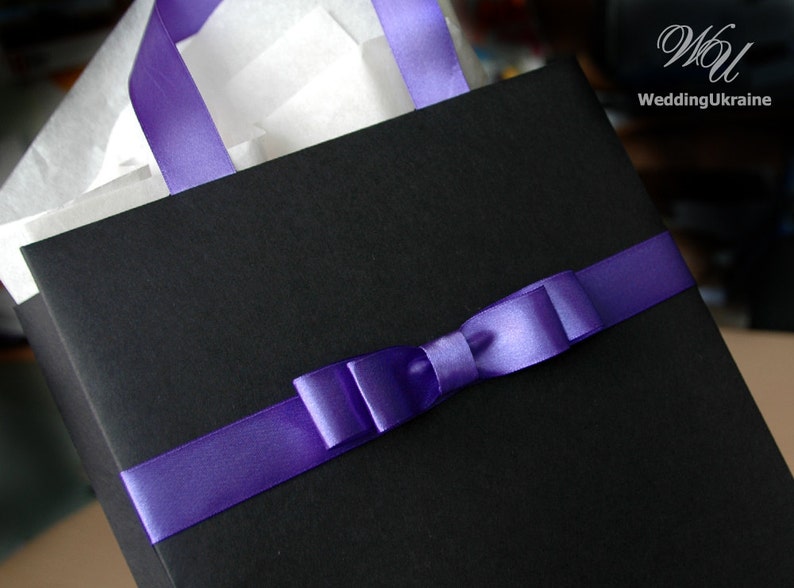 May include: A black gift bag with a purple satin ribbon tied in a bow. The bag is filled with white tissue paper.
