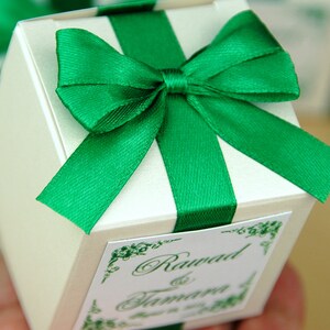 May include: A white favor box with a green satin ribbon and bow. The box has a label with the text "Rawad & Tamara" in green cursive.