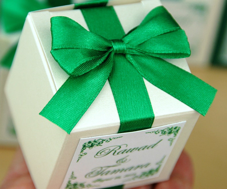 May include: A white favor box with a green satin ribbon and bow. The box has a label with the text "Rawad & Tamara" in green cursive.