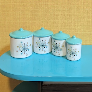 May include: Four white ceramic canisters with turquoise lids. Each canister features a colorful starburst design. The canisters are arranged on a turquoise surface against a yellow background.
