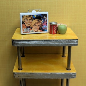 May include: A miniature scene featuring a white lunchbox with "Hardy Boys Mysteries" graphics, a small red cola can, and a green apple. The items are arranged on a yellow table with silver trim, evoking a nostalgic, vintage aesthetic.