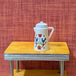 May include: A small, white ceramic pitcher with a lid, handle, and spout. The pitcher is decorated with blue birds, red flowers, and green leaves. It sits on a small, yellow table with silver trim and legs.