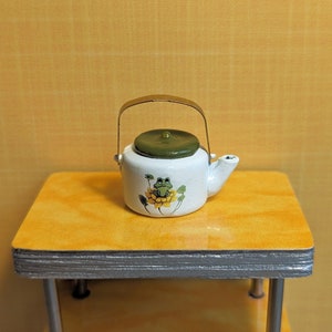 May include: A small white ceramic teapot with a green lid and a gold handle. The teapot features a frog design on a yellow flower. The teapot is sitting on a yellow table with silver legs.