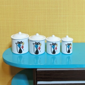 Może przedstawiać: Zestaw czterech białych ceramicznych pojemników z pokrywkami, każdy ozdobiony czarną sylwetką kota i geometrycznym wzorem w stylu retro w kolorach czerwonym, niebieskim i żółtym. Pojemniki ustawione są na turkusowej powierzchni.