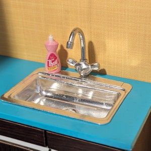 May include: Miniature kitchen sink with a chrome faucet and a pink bottle of "Thrill" dish soap. The sink is set on a blue countertop with a yellow wall in the background. A charming, vintage-style kitchen scene.