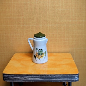May include: A white ceramic pitcher with a green lid and handle. The pitcher features a cartoon frog on a yellow flower. The pitcher is on a small, orange table with a yellow background.