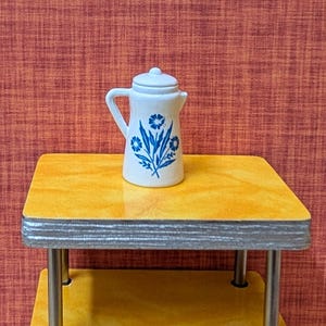 May include: A small, white ceramic pitcher with a lid, adorned with blue floral designs. The pitcher sits on a yellow table with silver trim and legs. The background is a textured, reddish-orange color.