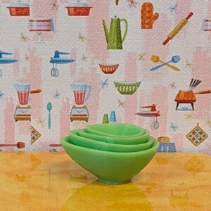 May include: A set of four nested green bowls. The bowls are made of a solid material and have a smooth, rounded shape. The background features a patterned wallpaper with kitchen-themed illustrations.