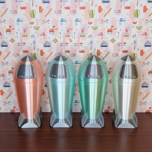 May include: Four retro-style trash cans in shades of copper, teal, green, and silver. Each can is shaped like a rocket with a silver lid and base. The word "TRASH" is printed on the front of each can. The background features a kitchen-themed wallpaper.