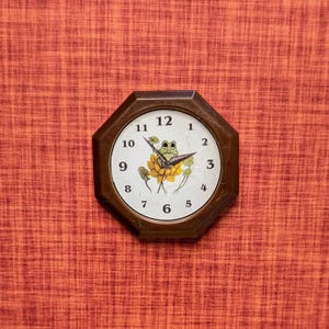 May include: A small, octagonal wooden clock with a white face. The clock features black numerals and hands, and a frog and flower design in the center. The clock is set against a textured orange background.