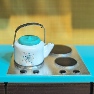 May include: A miniature white teapot with a teal lid and a silver handle sits on a silver stovetop with black burners. The teapot has a colorful starburst design. The stovetop is on a turquoise surface.