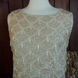 May include: A white beaded top with a fringe trim. The top is sleeveless and has a scoop neck. The beads are arranged in a pattern of scallops.