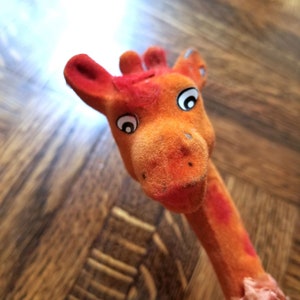 May include: Orange plush giraffe toy with black eyes and a pink ribbon around its neck. The giraffe is lying on a wooden surface.