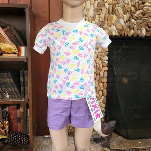 May include: A white short-sleeved shirt with a colorful floral print and a purple pair of shorts. The shirt has a white collar and a white trim around the sleeves. The shorts have a waistband with a small pink tag that says "PIXIE" in pink letters.