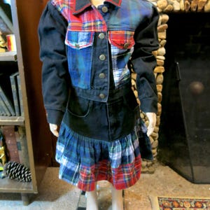 May include: A black denim jacket with a plaid patchwork design. The jacket has a button closure and a collar. The jacket is paired with a black denim skirt with a plaid ruffle trim.