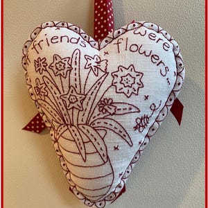 May include: A white heart-shaped pillow with red stitching and a red ribbon. The pillow is embroidered with a bouquet of flowers and the text "Friends were flowers".