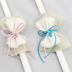 Könnte beinhalten: Zwei weiße Kerzen mit dekorativen Schleifen. Eine Schleife ist rosa und die andere blau. Beide Schleifen sind aus Jute und haben einen kleinen Kreuzanhänger.