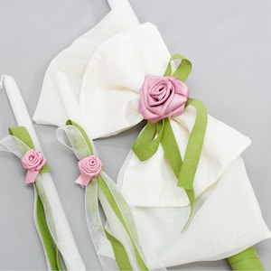 Könnte beinhalten: Zwei weiße Kerzen mit grünen und durchsichtigen Schleifen und rosa Rosenakzenten. Die Kerzen sind mit einer Satinrose und einem grünen Band verziert.