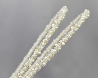 May include: Two white pearl beaded headbands, twisted together, on a gray background.